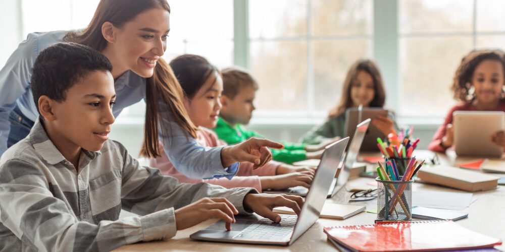 Teacher Teaching School Kids Using Laptop In Classroom.