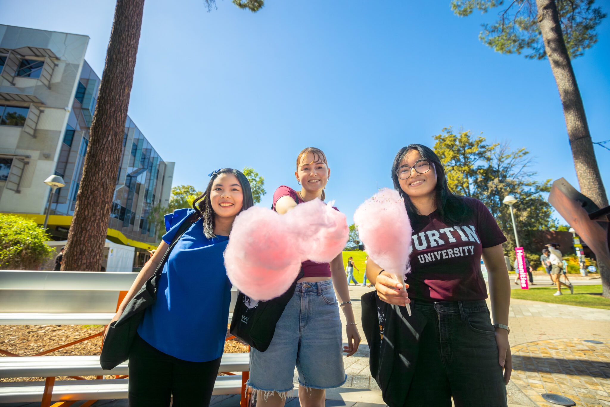 Three people outdoors on campus holding large pink fairy floss, with university buildings and trees in the background.