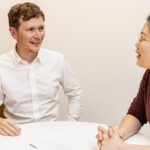 Two people seated at a small table in an office, having a face‑to‑face conversation with papers and a pen between them.