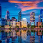 City skyline at dusk with illuminated high‑rise buildings reflected on calm water under a colourful evening sky.