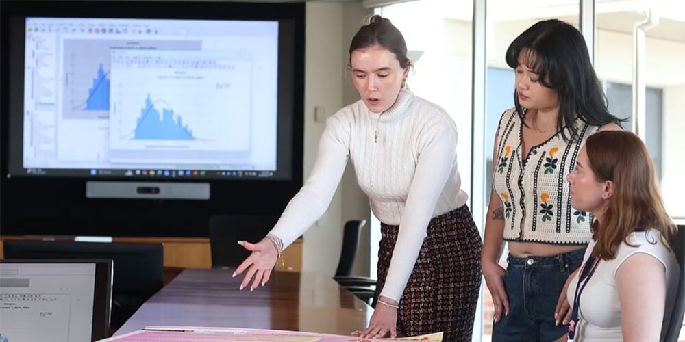 Woman in white pointing at information sheet on table with screen in the background and two women to her right.