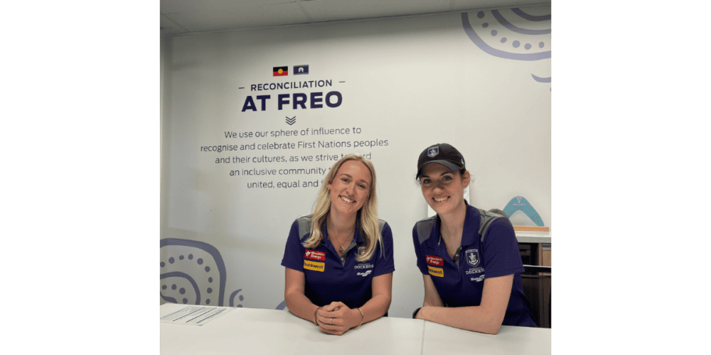 Two people in Fremantle Dockers shirts seated at a table in front of a Reconciliation at Freo sign.