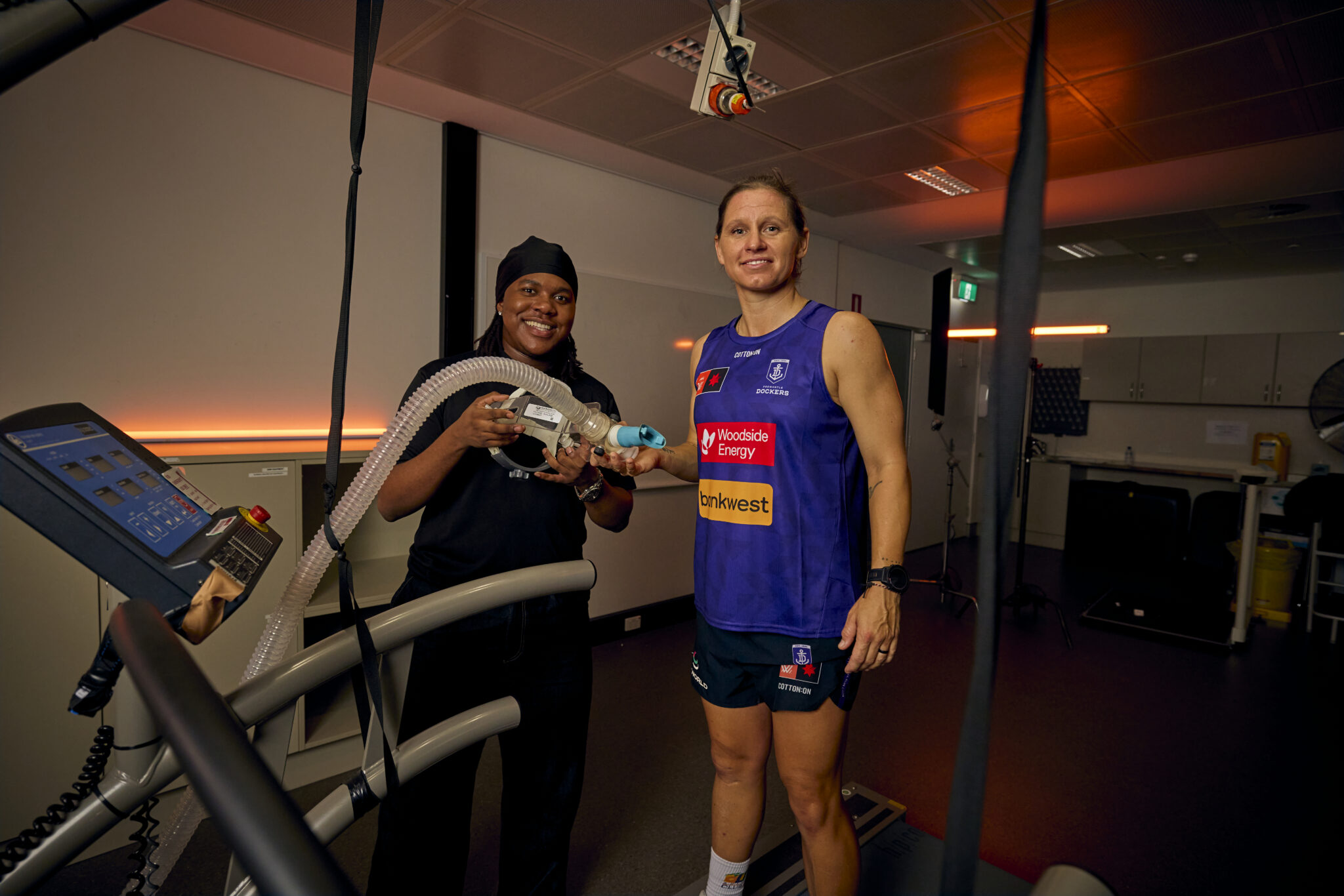 Two people holding exercise testing equipment beside a treadmill in a sports science lab.