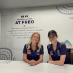 Two people in Fremantle Dockers polo shirts seated at a table in front of a Reconciliation at Freo sign.