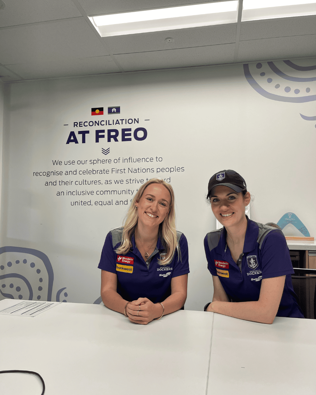 Two people in Fremantle Dockers polo shirts seated at a table in front of a Reconciliation at Freo sign.