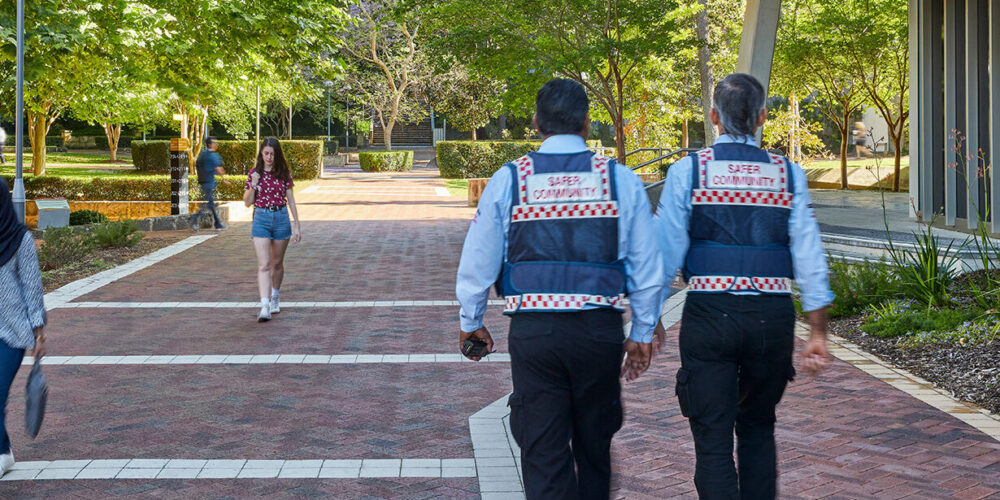 2 Safer Community officers walking next to students. The officer's backs are facing the reader.