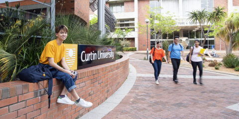Student sitting on wall next to a sign that says Curtin University