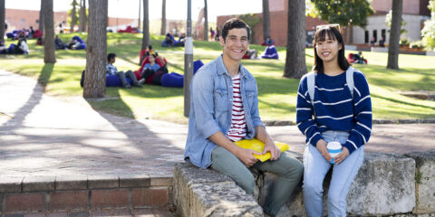 Two students sitting on a wall outside smiling at camera