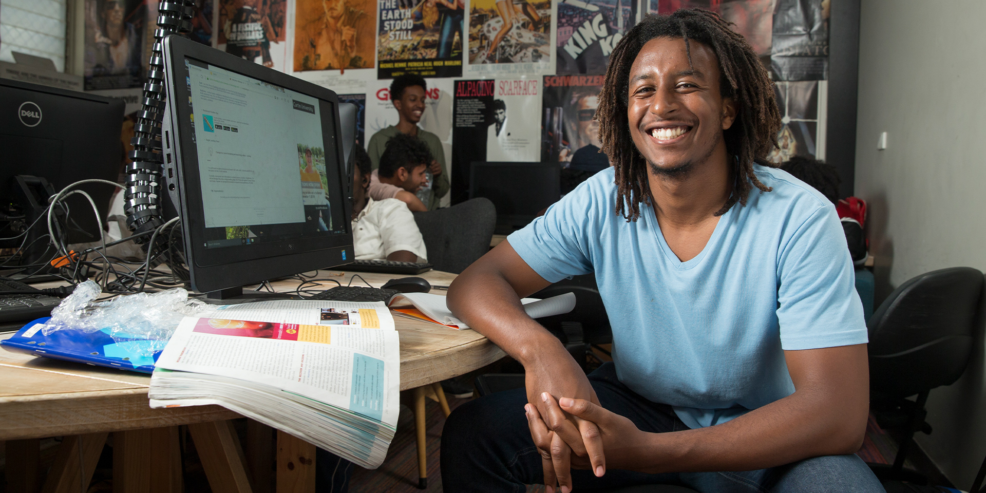 Student wearing a blue t-shirt smiles next to a computer