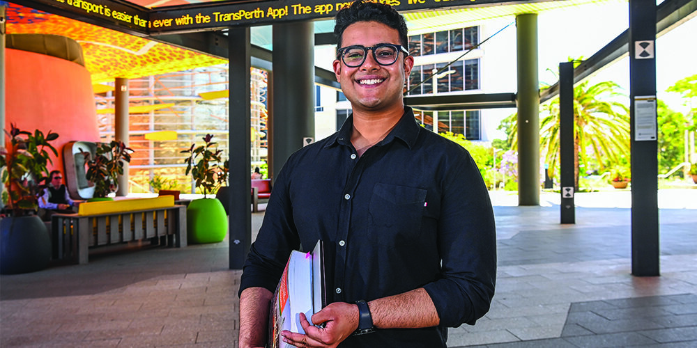 A male student wearing a black shirt smiles holding textbooks under his arm