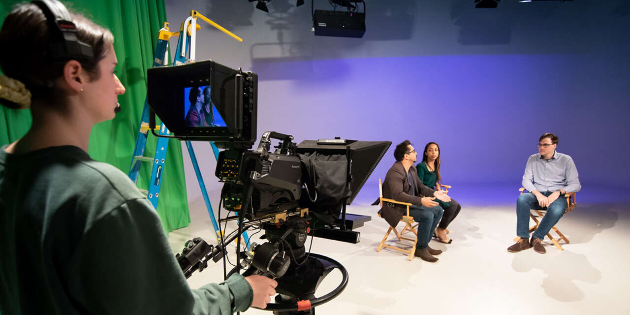 Three students sitting on a film set being filmed by a student behind a camera.