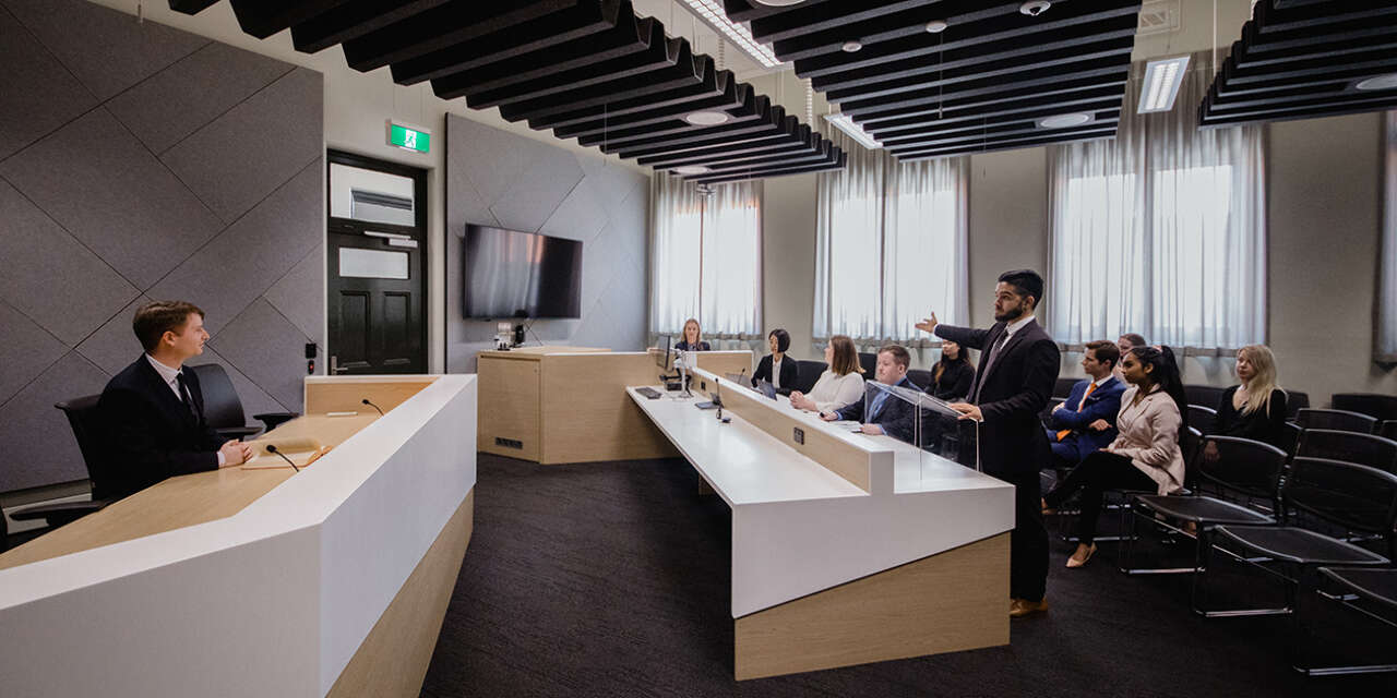 A moot court setting with judge sitting at a desk facing another desk where a man stands. Behind him are audience seats, where several people sit and watch.