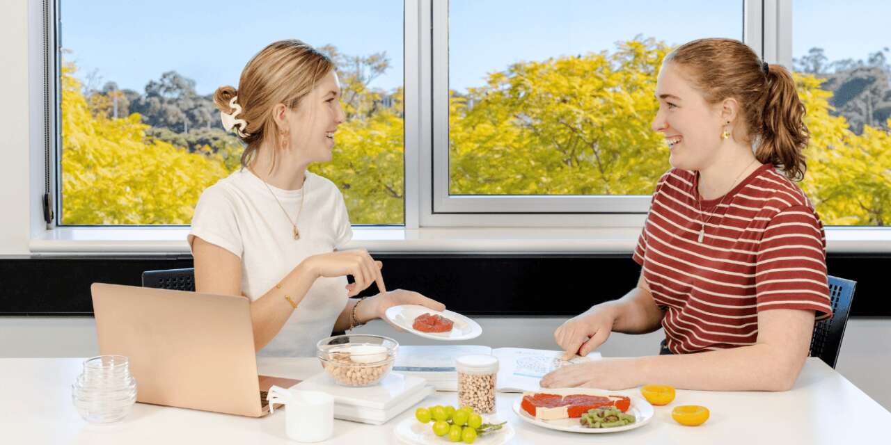 Two Nutrition students sitting at a table, smiling at each other, with an assortment of fruit and meat displayed on the table.