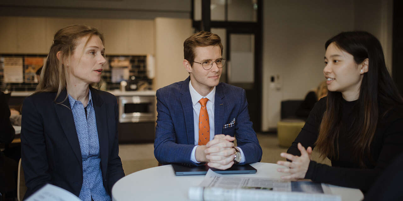 Three professionals sit around a table engaged in a conversation.