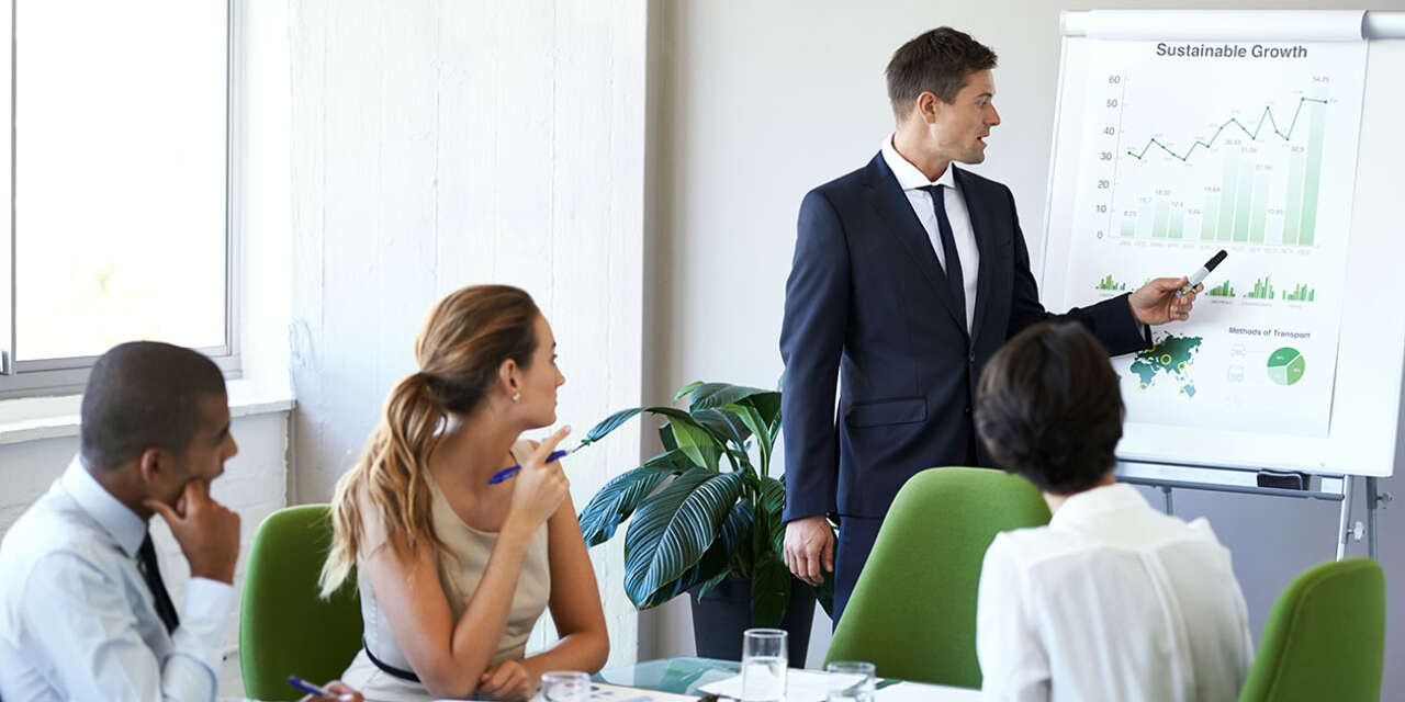 Man in business attire pointing at a chart labelled 'business growth' while a table of colleagues looks on.