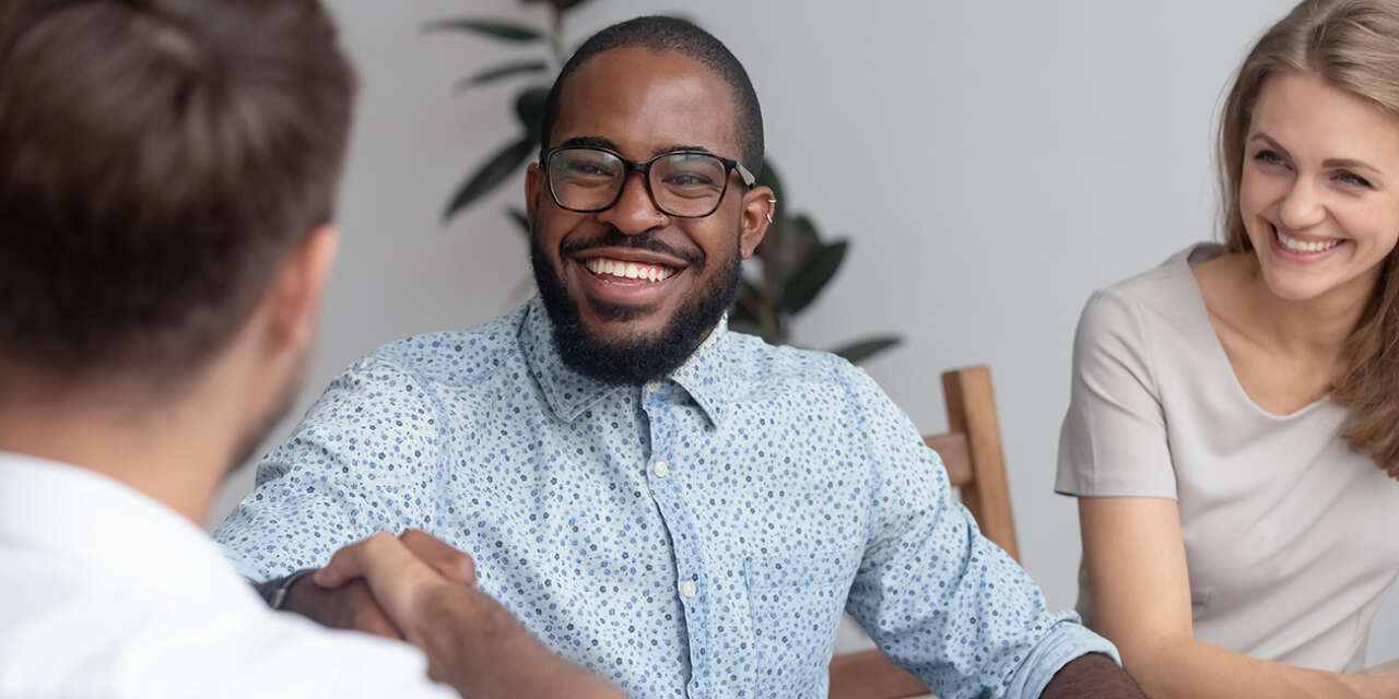 Three people engaging in conversation in a professional setting. The focus is on a male-presenting person with dark brown skin, short black hair and beard, black glasses and a light blue business shirt. He has a broad smile. A female-presenting woman with long blonde hair and a light top smiles next to him, and a male presenting person with short brown hair faces him and is only visible from the back.