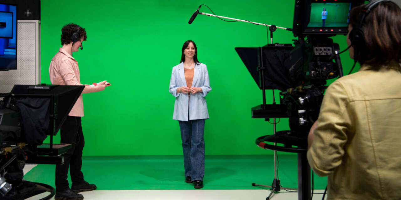 A female in her 20s stands in front of a green screen. A boom mike is positioned above her, and in the foreground another person is viewing her through a professional camera.