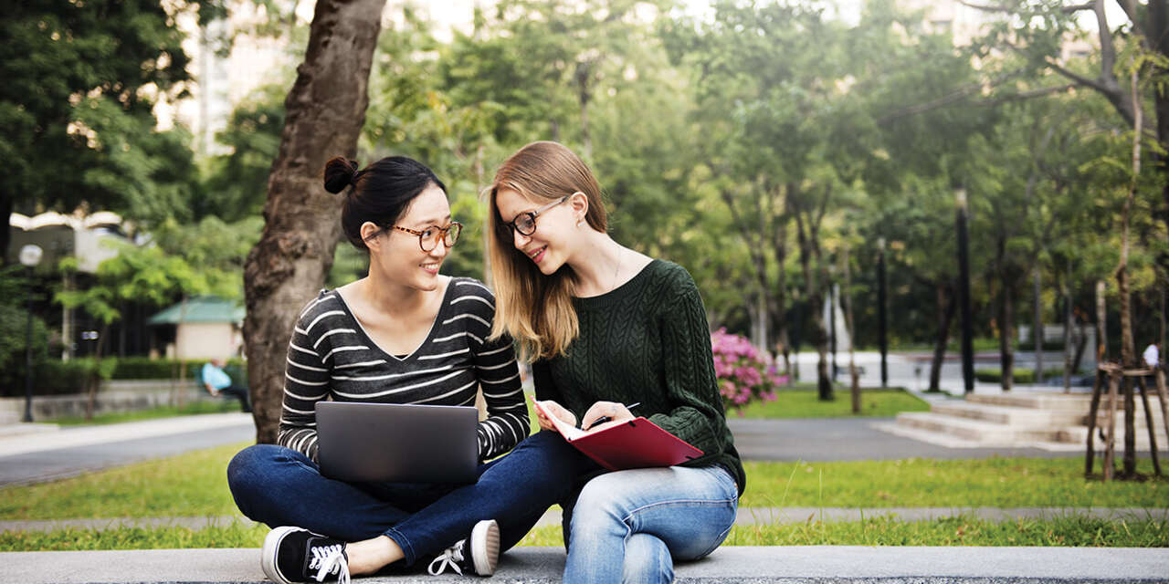 Two female students sitting down smiling and looking at a laptop and book.