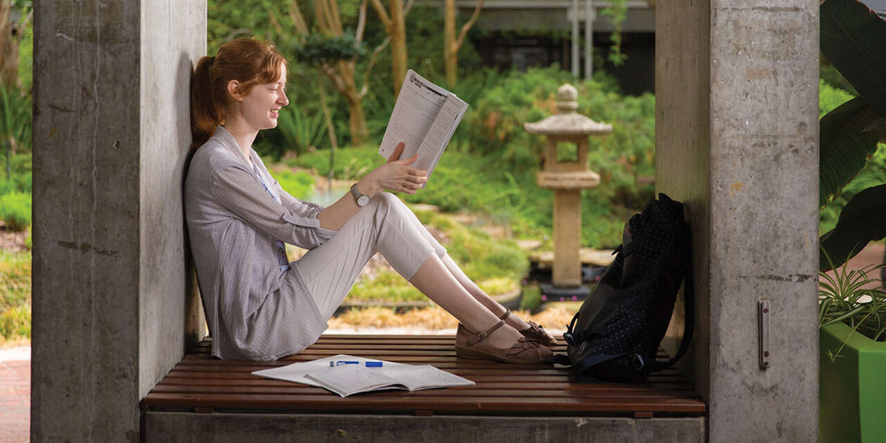 A student smiling and reading a book with greenery in the background.