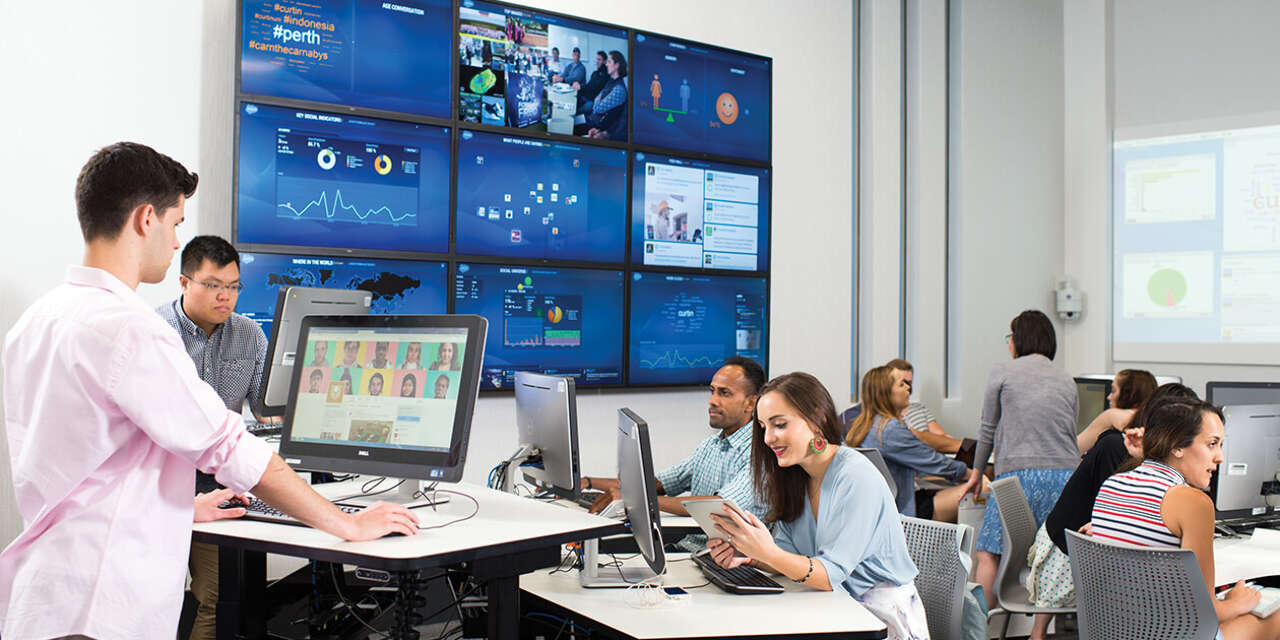 Students working on computers with large screens showcasing different sets of data in the background.