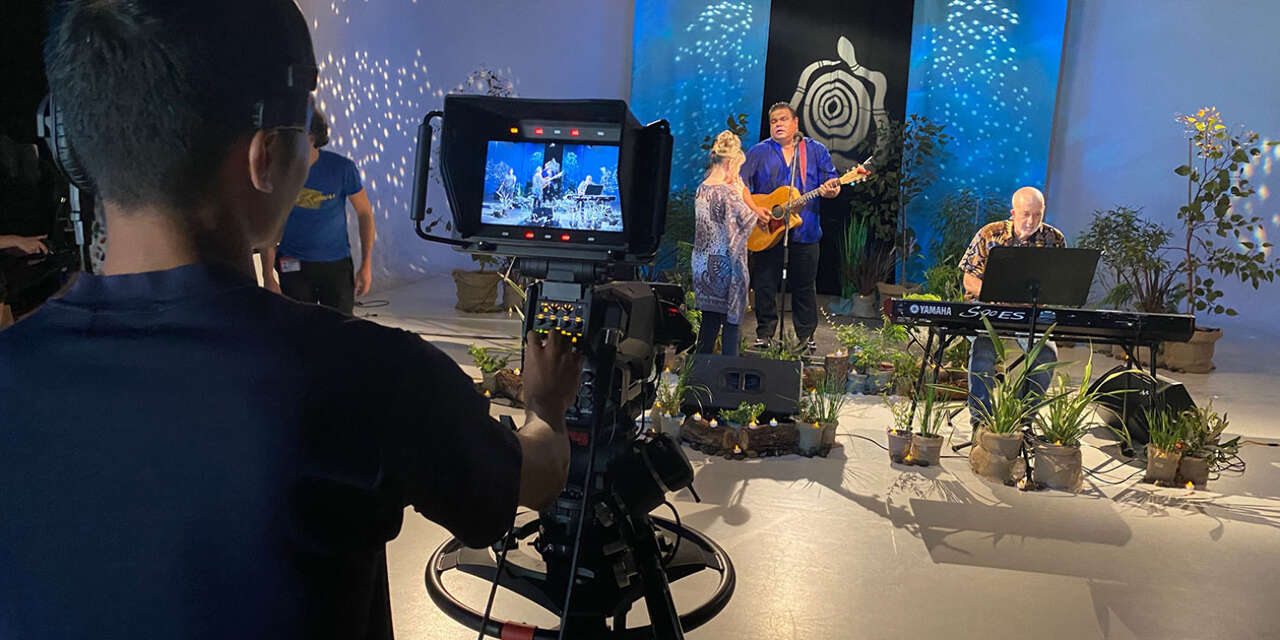 Person playing guitar on a studio set, surrounded by lights and cameras