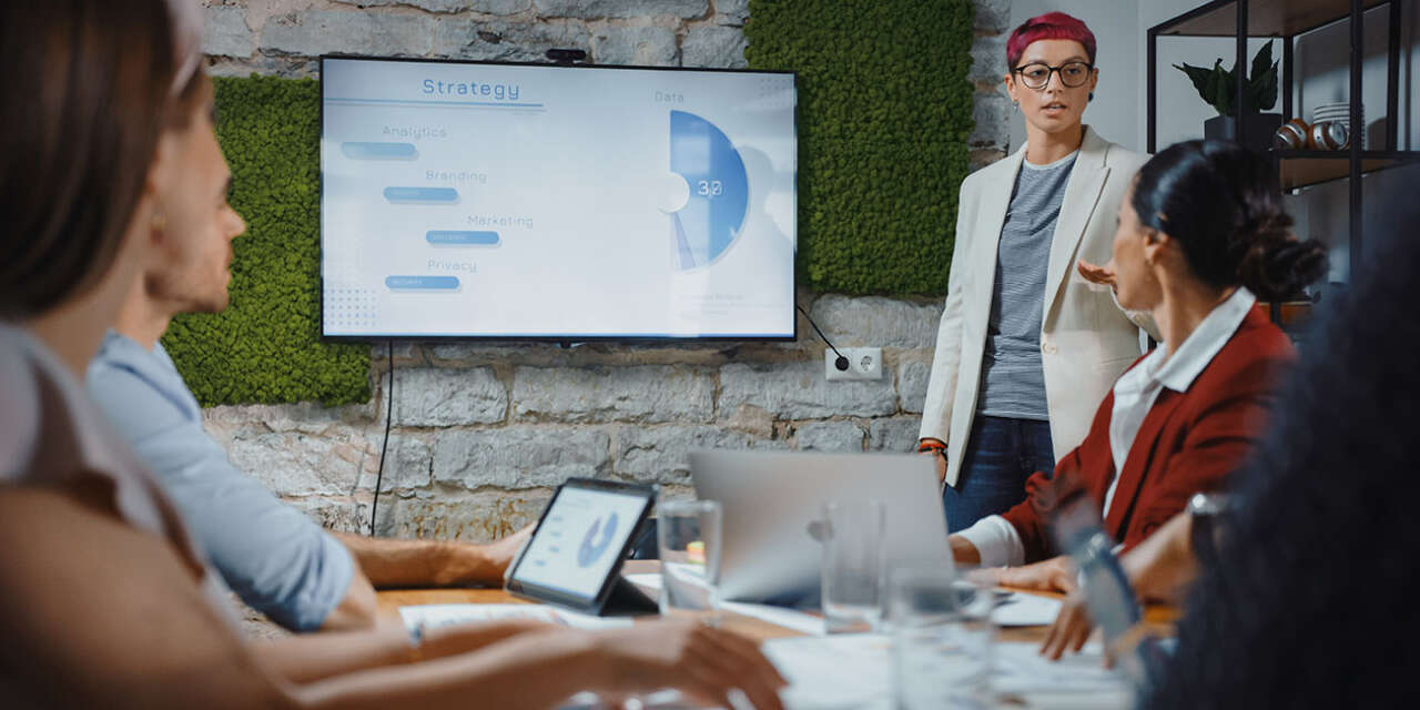 Group of people sitting around a table looking at a presenter and a screen.