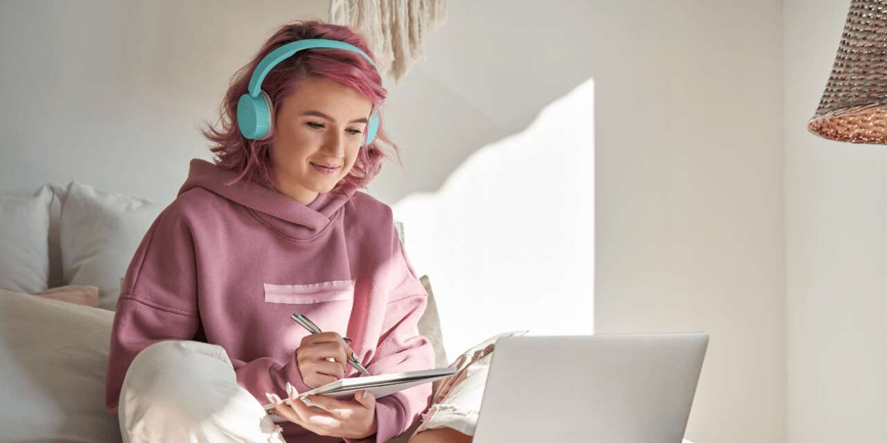 Woman with pink hair writes in a notepad while looking at her laptop. She is sitting on her bad.