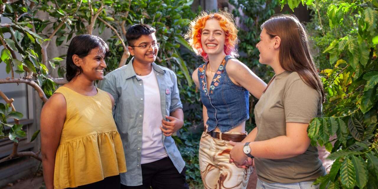 Four people stand outside in a group, chatting. The group is of three female-presenting people and one male-presenting person with different ethnicities. A women with white skin and brilliant orange hair looks directly to camera with an inviting grin.