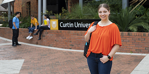 A student in an orange shirt, looking at camera and smiling, standing in front of the Curtin sign