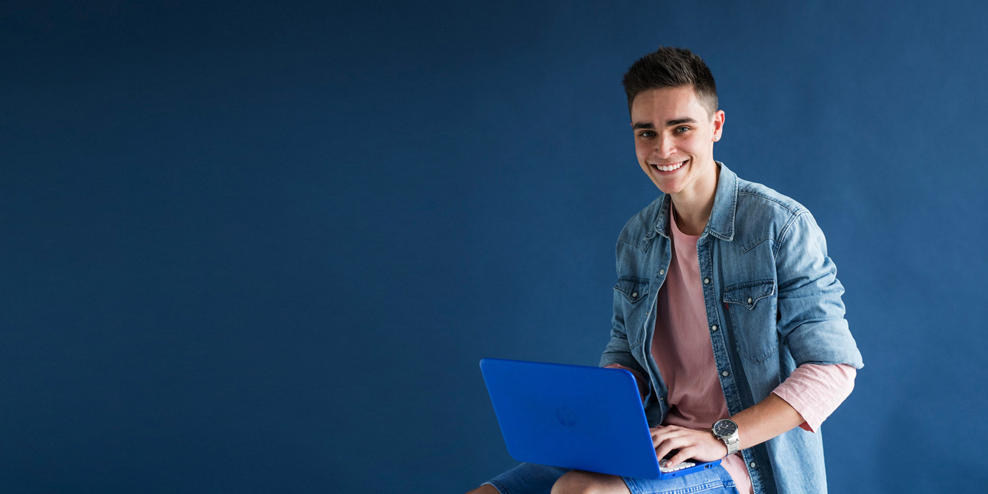 A young male sitting on a stool in front of a blue background, Holding a laptop